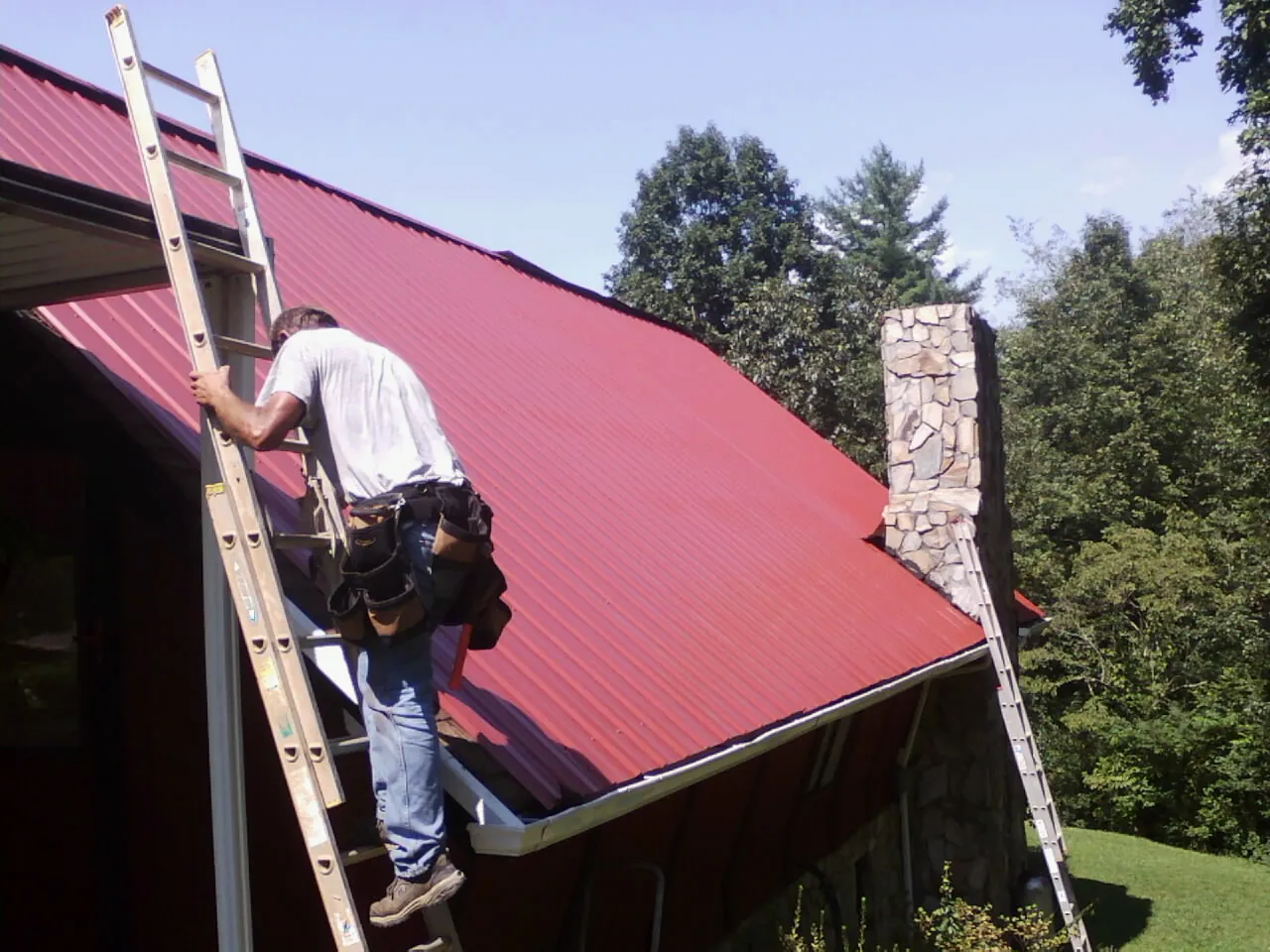 Roofer on a ladder beside a red metal roof and stone chimney — Soffit & Fascia Repair by Roofing Brothers in Brookfield